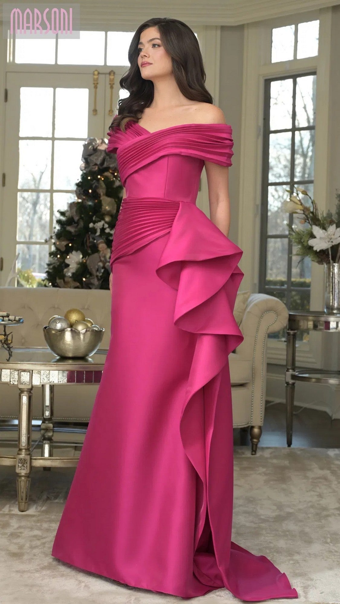 Indoors, a woman wears the Marsoni MV1354 long formal dress in fuchsia—an off-shoulder, floor-length mermaid gown featuring draped and pleated details.